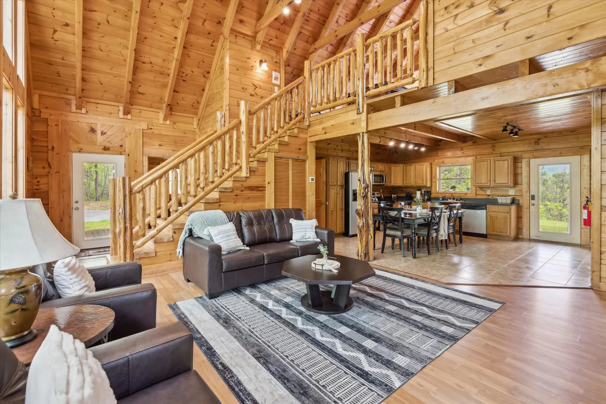 An open-plan living space that integrates a dining table with chairs, a wooden staircase, a sofa with throw pillows, and large windows. The layout encourages socializing, casual dining, and connection to the living and game rooms.