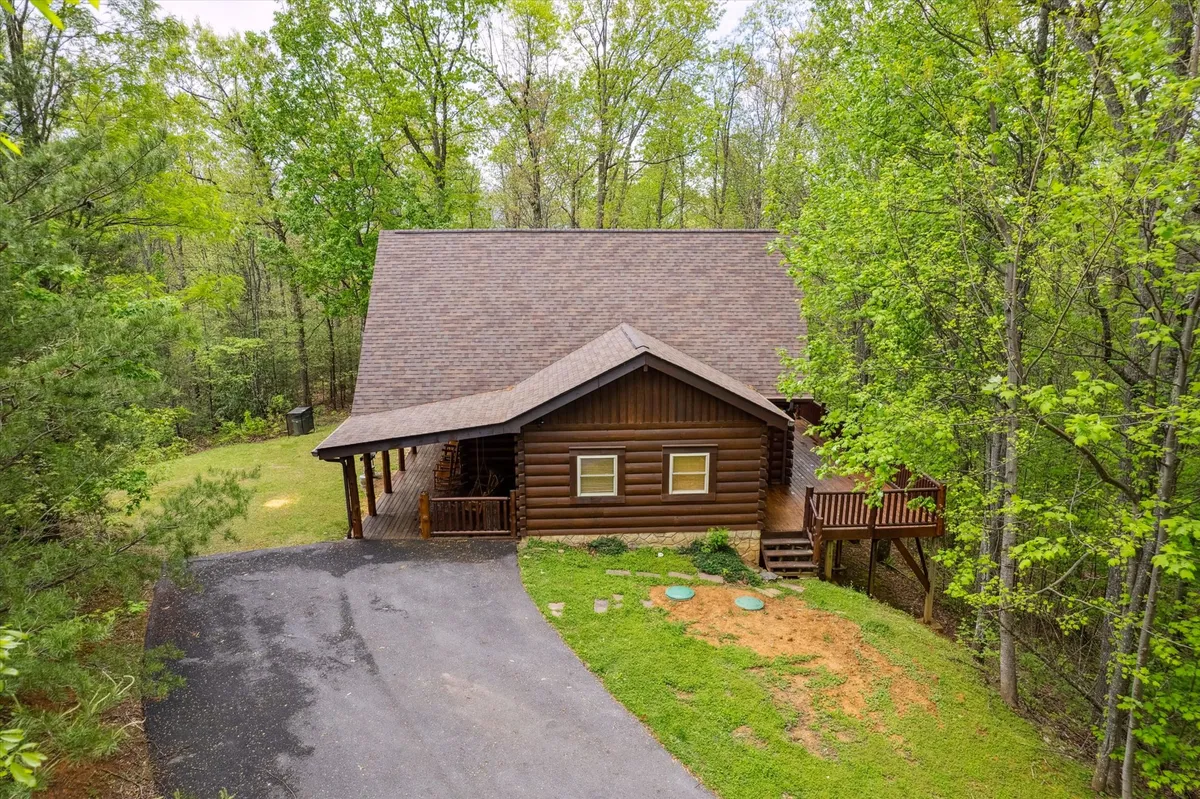 This exterior shot captures the log cabin structure nestled among lush greenery, with a driveway leading up to the entrance. The inviting porch provides a glimpse of a peaceful mountain setting, ideal for unwinding after outdoor adventures.