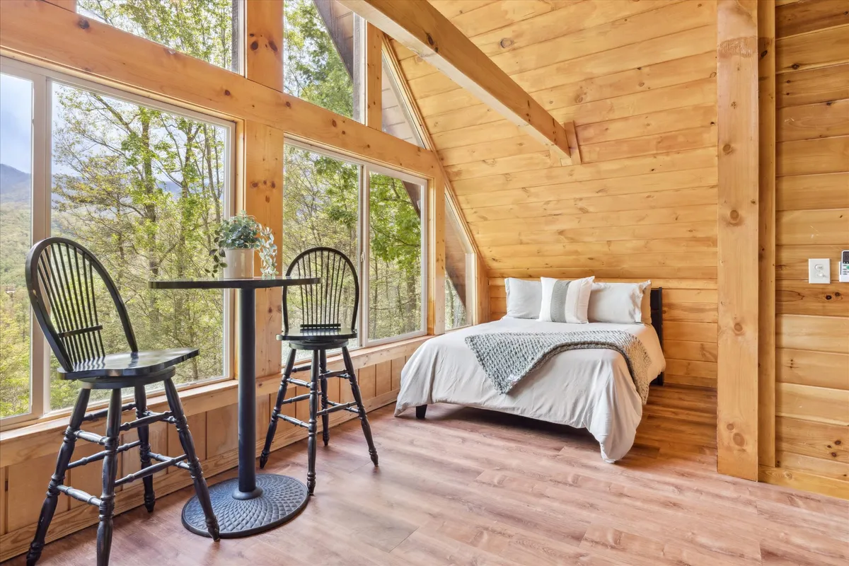 A serene room with a soft-linen bed positioned near large windows that offer panoramic views of the surrounding landscape. A small table with chairs by the window invites moments of reflection, reading, or morning coffee while taking in the scenery.