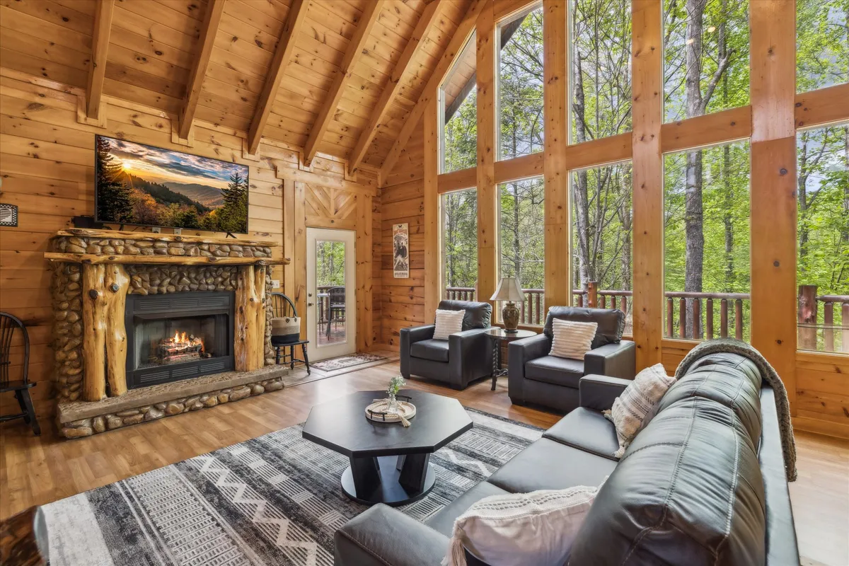 A spacious living room with large windows framing the forest view, a stone fireplace as a focal point, a comfortable seating arrangement with sofas, and a TV mounted on the wall. Natural light fills the room, creating a bright and welcoming space for gatherings.