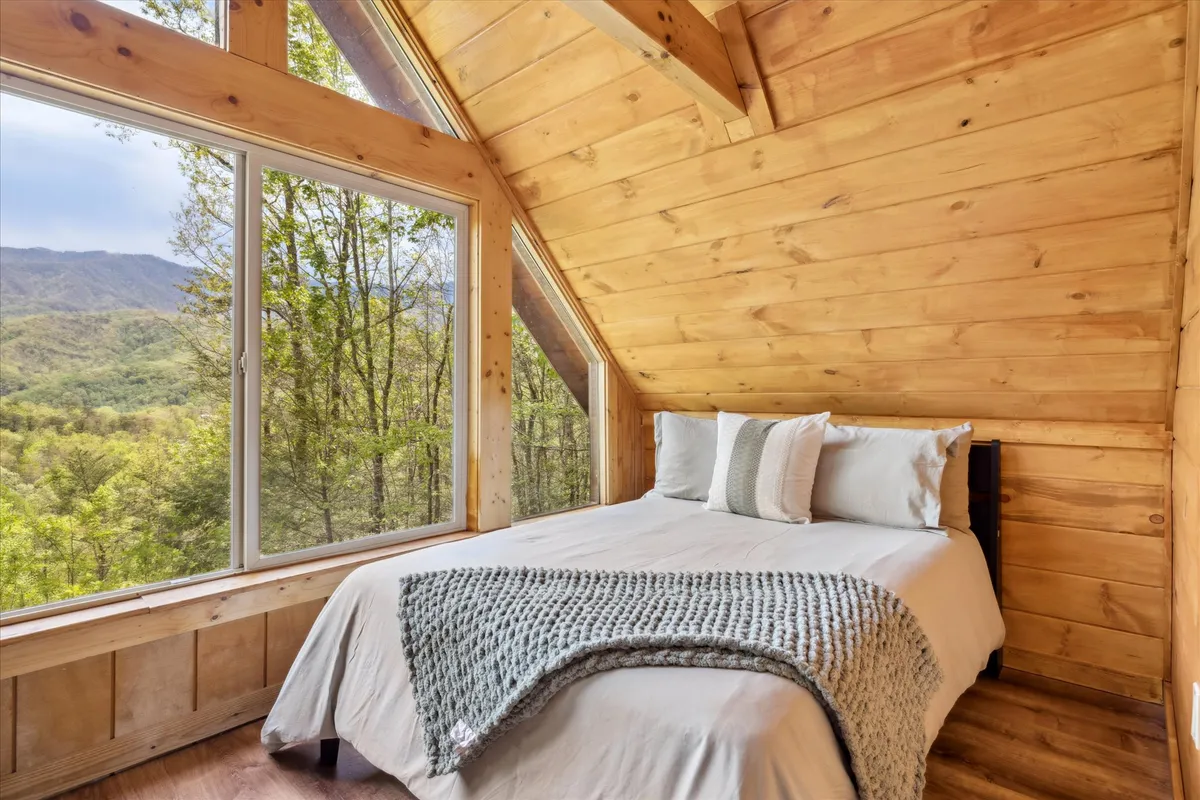 A cozy bedroom with large windows framing a stunning mountain view. The bed is adorned with decorative pillows and a soft blanket, and the space is surrounded by wood-paneled walls and ceiling, creating a warm, tranquil atmosphere. Natural light floods the room, enhancing the serene mood.